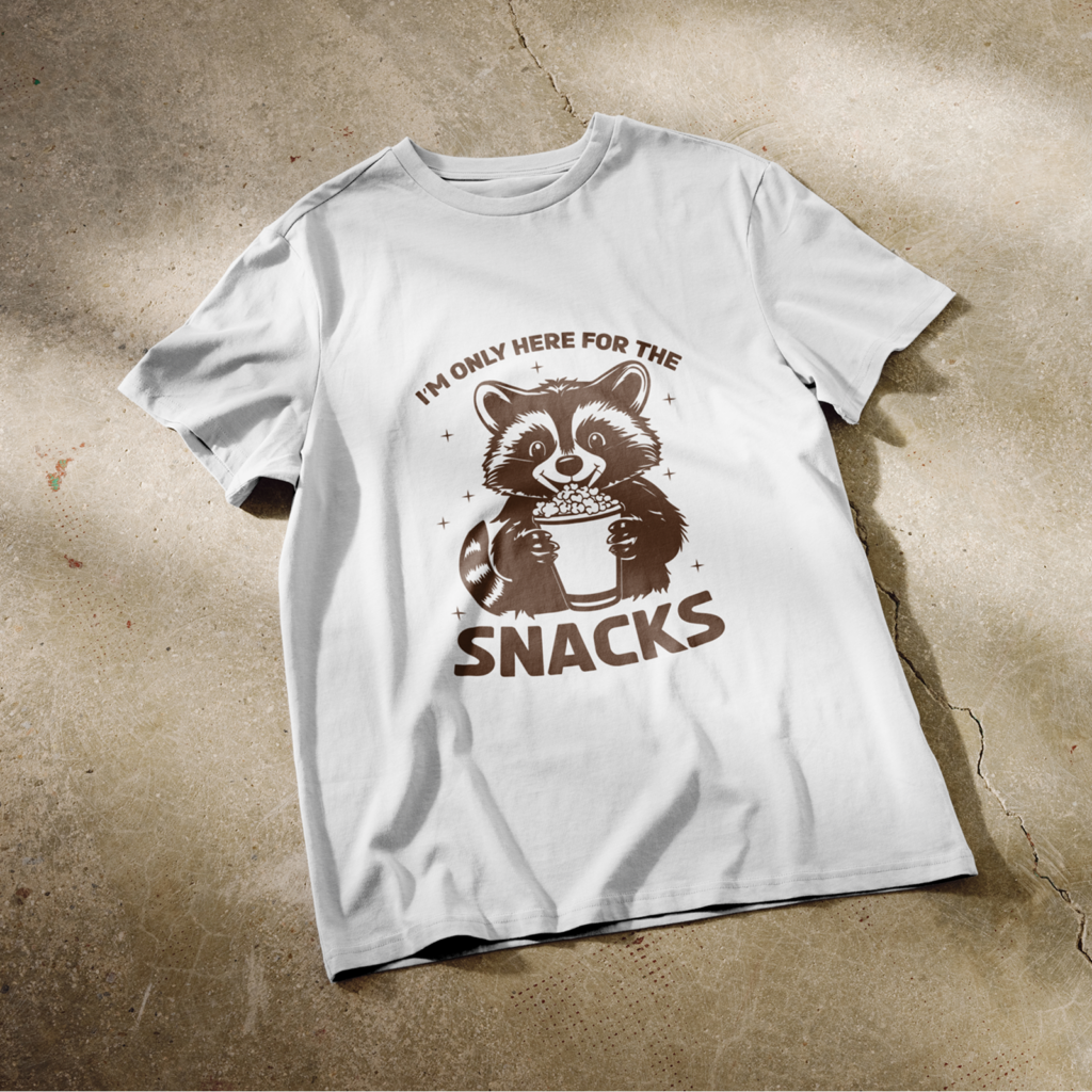 White t-shirt on concrete floor, featuring a cute raccoon holding popcorn with text "I'm Only Here for the Snacks".