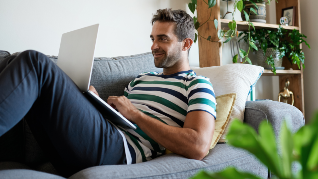 A man sitting on a couch, focused on his laptop, with a relaxed expression in a cozy living room setting.