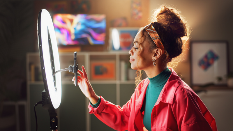 A woman with curly hair in a bun, wearing a pink jacket and headband, adjusts her phone on a tripod with a ring light.