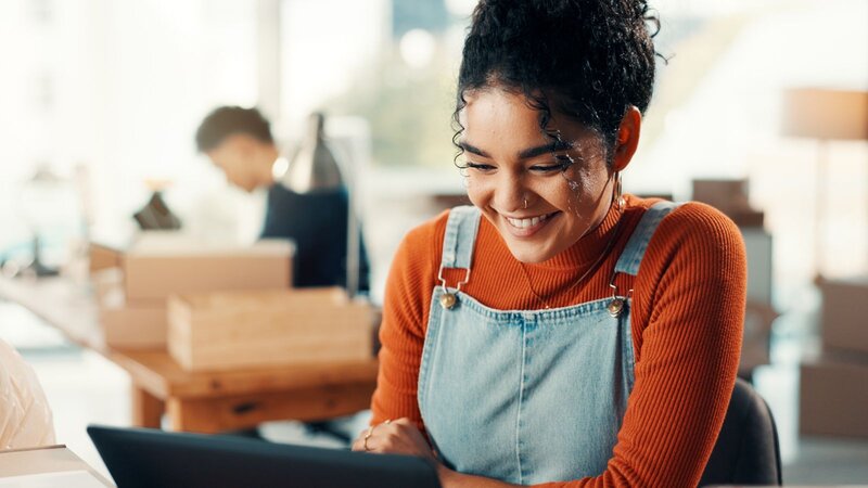 A young woman with curly hair, smiling while sitting at a desk with a laptop, wearing an orange sweater and blue overalls.