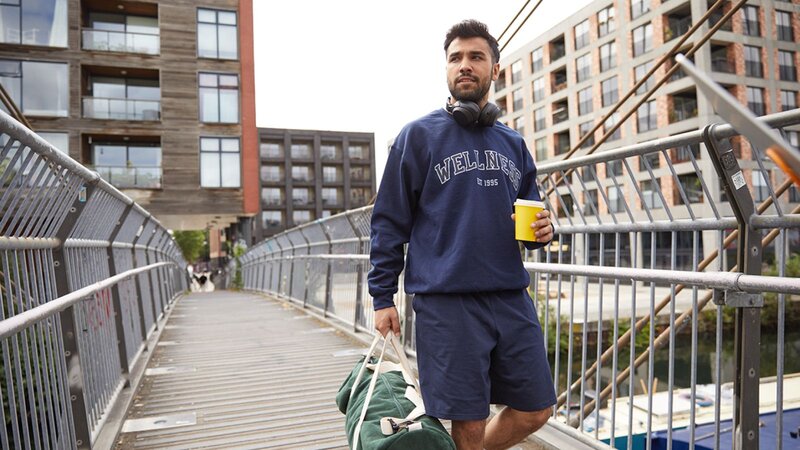 A man walking on a bridge with a drink in one hand and a duffel bag in the other, wearing a navy sweatshirt and shorts.