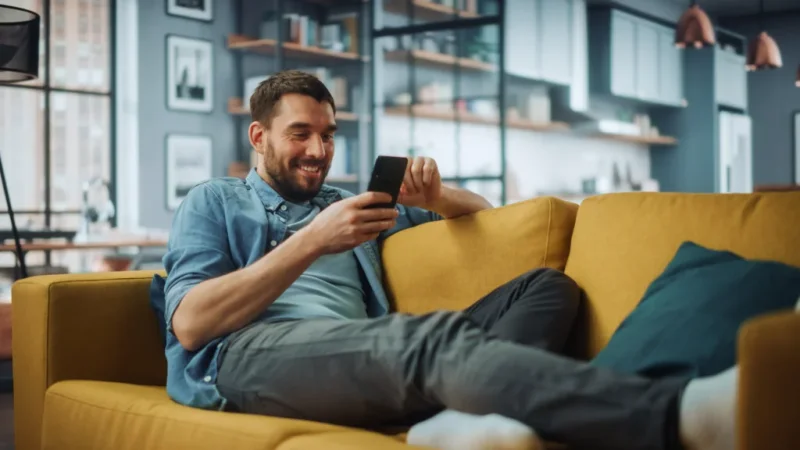 Man relaxing on a yellow sofa in a modern living room, smiling while looking at his phone. The room is with shelves and framed photos.