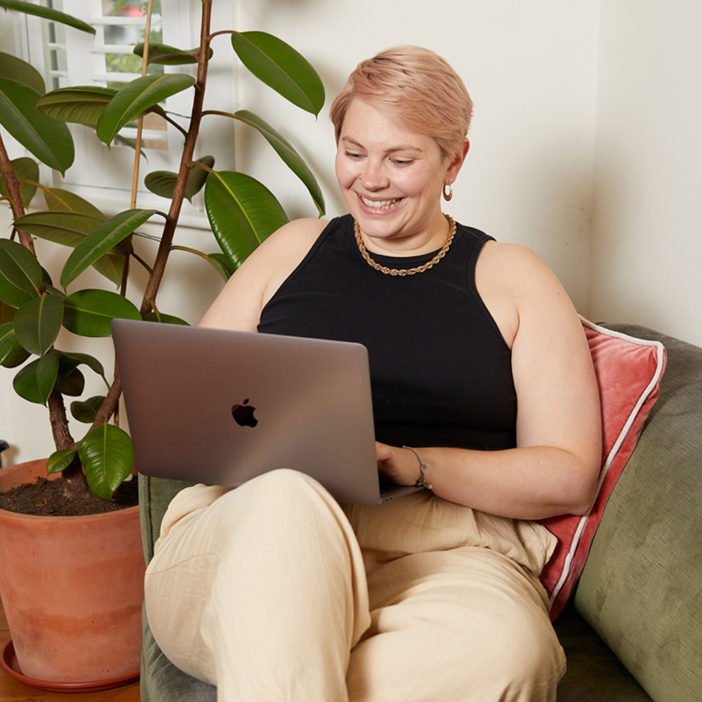 Woman with short hair sitting on a couch, smiling at a laptop. She's wearing a black tank top and beige pants, with a potted plant nearby.