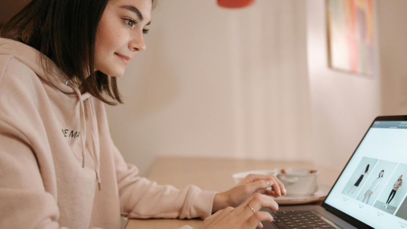 A woman in a camel hoodie sitting at a desk and using a laptop.