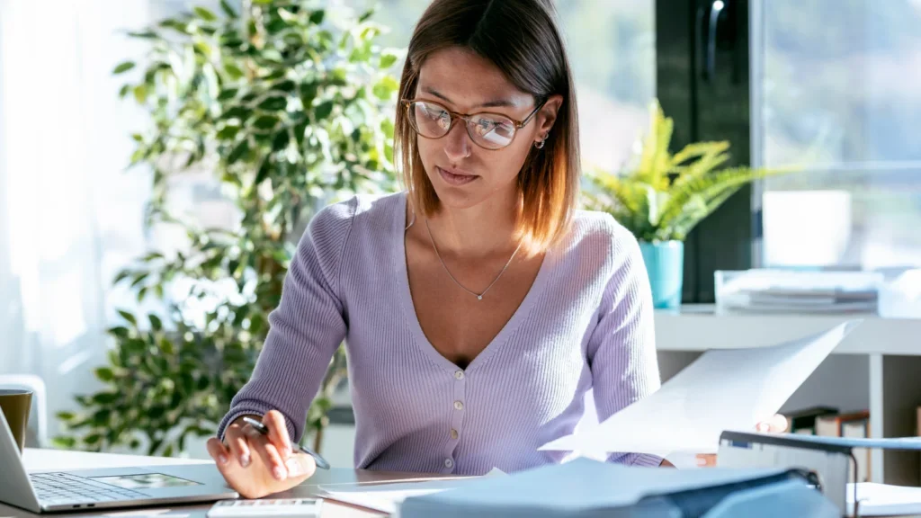 A woman in glasses is sitting at an office desk, working with papers and using a calculator. A laptop is nearby.