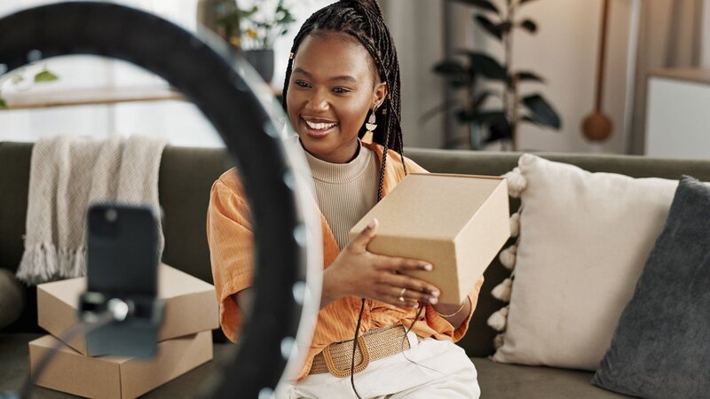 A woman in a cozy living room sits on a sofa, smiling as she presents a product in a cardboard box and records it with a smartphone.
