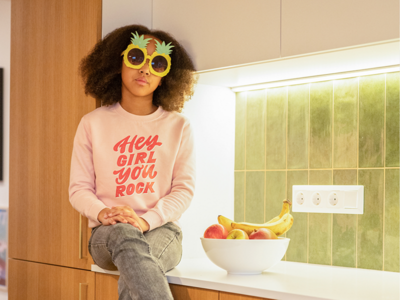 A young girl in pineapple sunglasses and a pink sweatshirt that says "Hey girl you rock" sits on a counter, showcasing her fun personality and style.