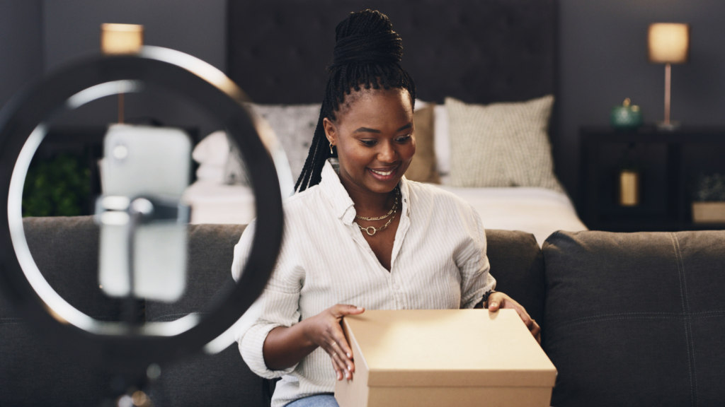 A woman is unpacking a delivery box during a livestream, using a smartphone on a stand with a ring light.
