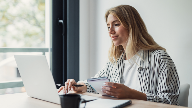 A smiling woman in a black-and-white striped blouse looks at a laptop screen while holding a pen and a notepad.