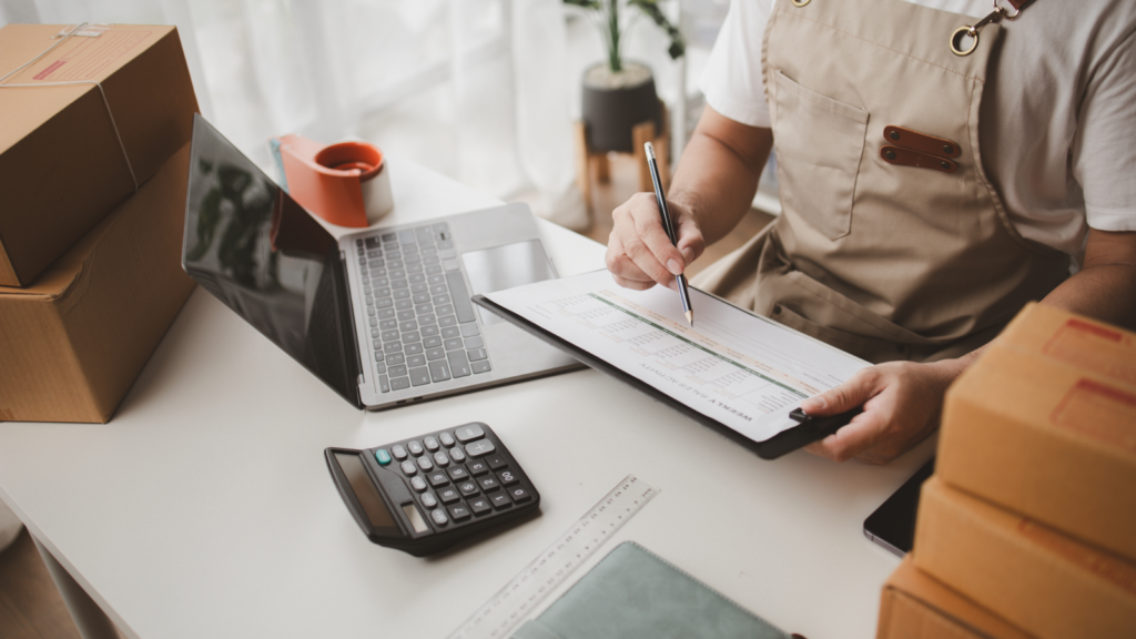 A man in an apron sits at a desk, focused on his laptop while surrounded by paperwork.
