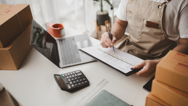 A man in an apron sits at a desk, focused on his laptop while surrounded by paperwork.