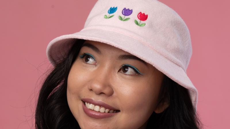 A woman smiling while wearing a pink bucket hat with flower embroidery, against a pink background.