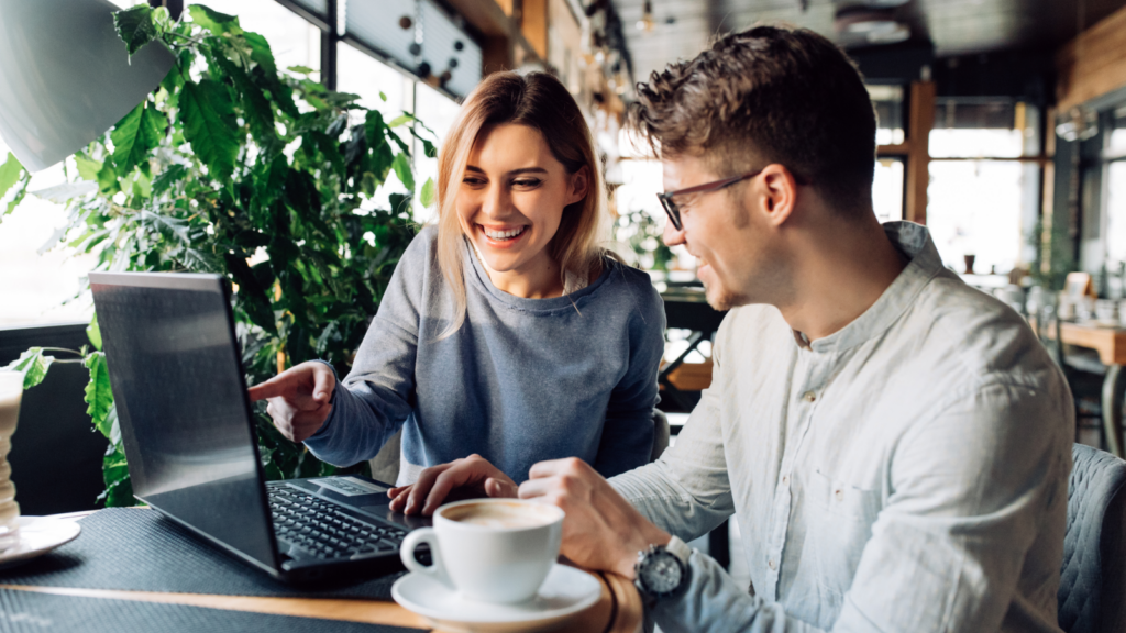 A smiling woman in a gray sweater points at a laptop screen, while a man in glasses and a white shirt sits beside her in a cafe.