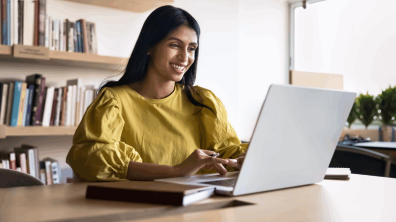 A woman in a yellow blouse smiles while using a laptop to find print-on-demand trends.