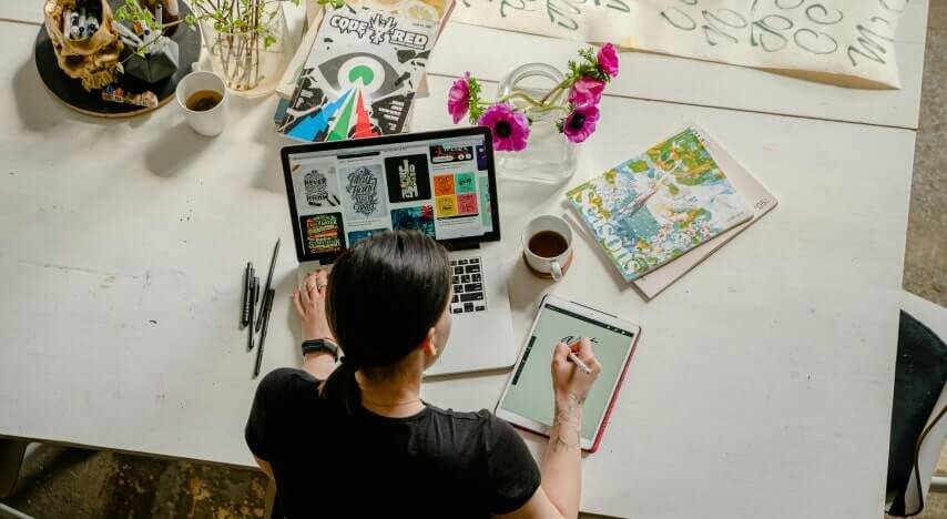 A woman drawing a design using a tablet while sitting on a desk with a laptop.
