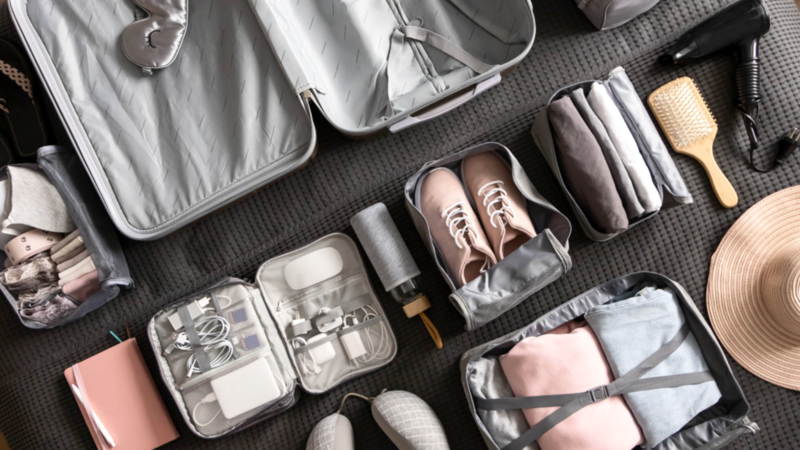 Open suitcase with neatly arranged items around: shoes, clothes, toiletries, tech gadgets, hairbrush, and a hat.