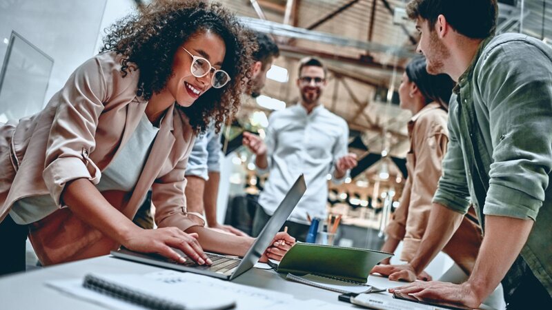 A smiling woman in glasses types on a laptop while a team of coworkers gathers around a table to plan a project in a bright industrial-style office.