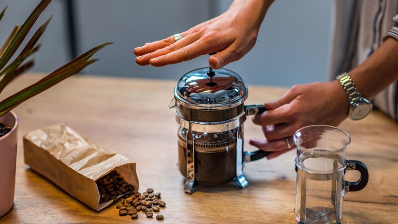 A person presses the plunger on a stainless steel French press next to a paper bag of spilled coffee beans and a glass cup on a wooden table.