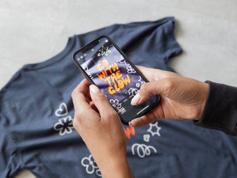 Hands hold a smartphone taking a picture of a “Go with the glow” t-shirt. The shirt has white floral doodles.