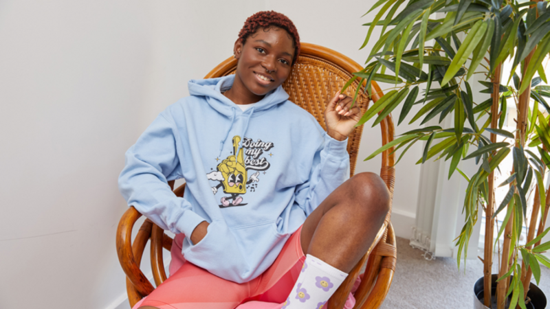 A smiling woman looks into the camera, wearing a blue “Doing my best” hoodie and pink yoga shorts.