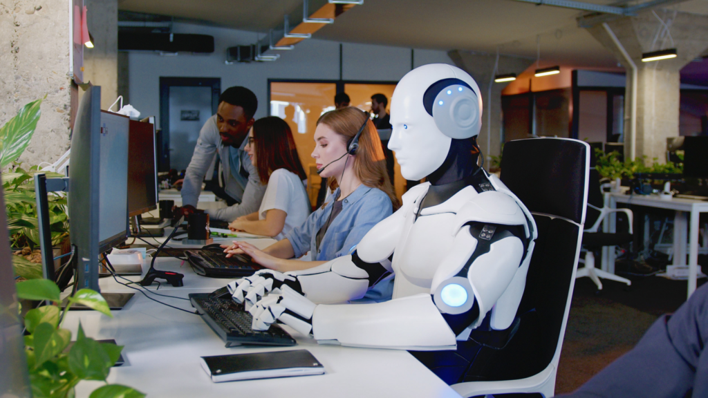 A robot seated at a desk in a modern office, surrounded by office supplies and a computer.
