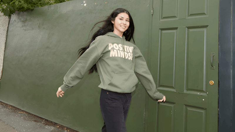 A smiling young woman walks down a small city street, wearing a dark-green “Positive Mindset” hoodie and black pants.