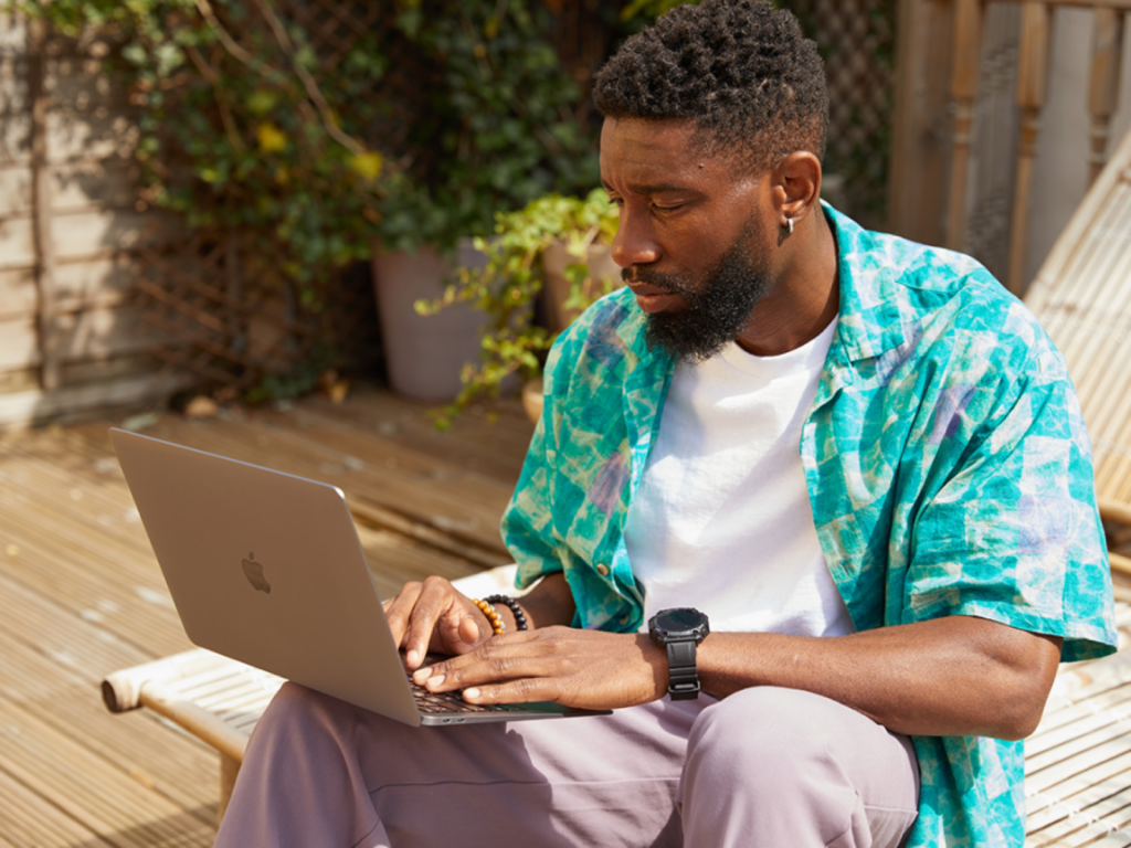 A man in a colorful shirt and casual pants works on a laptop outdoors on a wooden deck, surrounded by greenery.