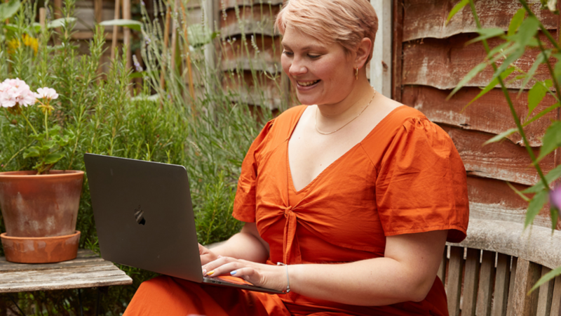 A smiling woman in an orange dress is sitting in a home garden with a laptop, searching for eco-friendly t-shirt printing options.