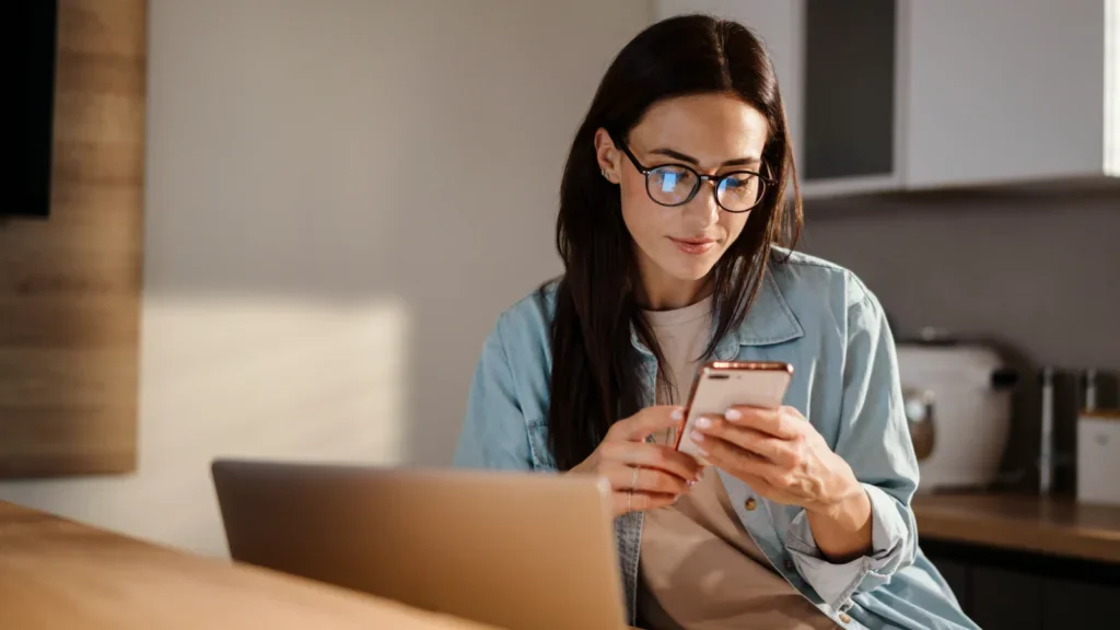 A woman wearing glasses is engaged with her phone and laptop, multitasking efficiently in a modern workspace.