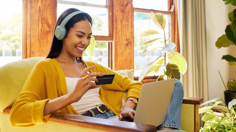 Smiling woman wearing headphones holding a credit card while shopping online on a laptop.
