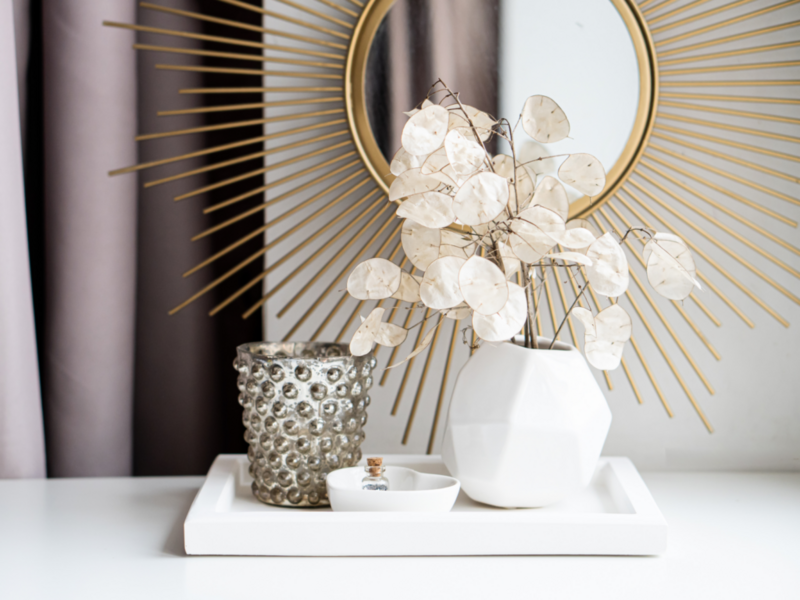 A white vase filled with flowers sits on a tray beside a stylish mirror, showcasing a trendy home decor setup.