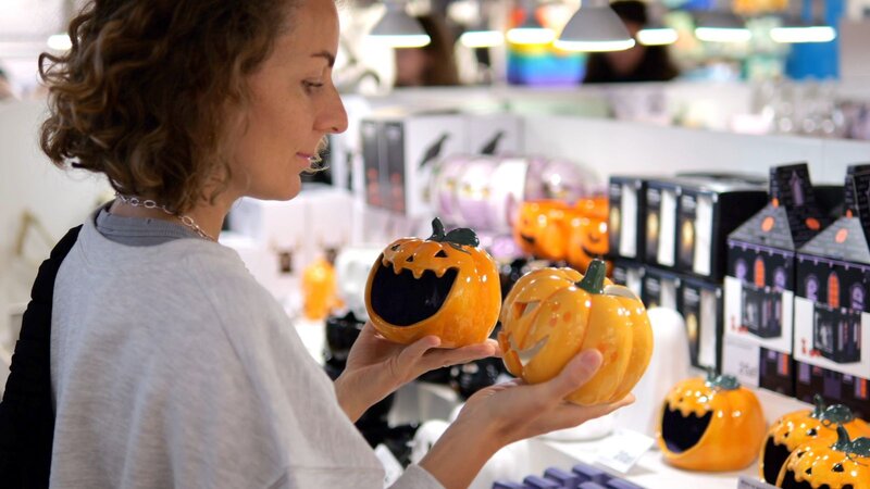 A shopper compares two ceramic jack-o'-lantern decorations in a store aisle stocked with Halloween home decor.