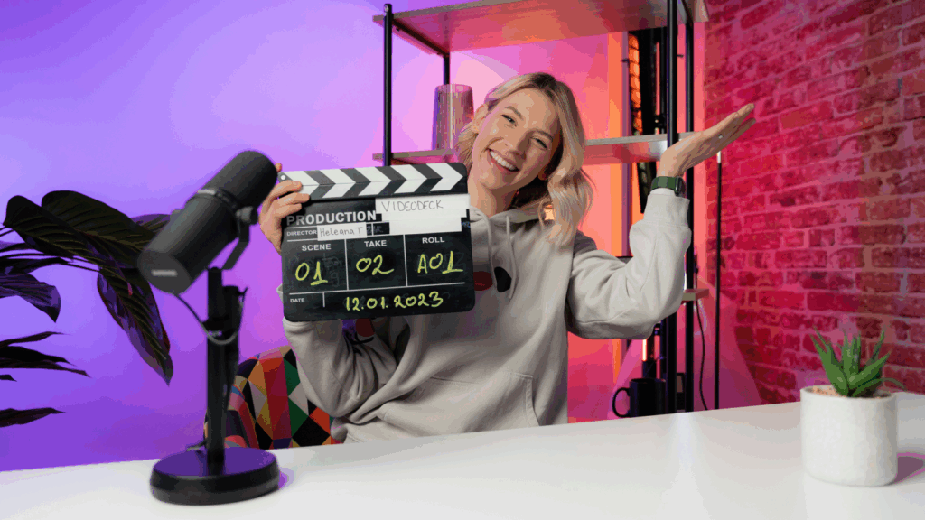 A smiling woman sitting in front of a microphone, holding a clapperboard.