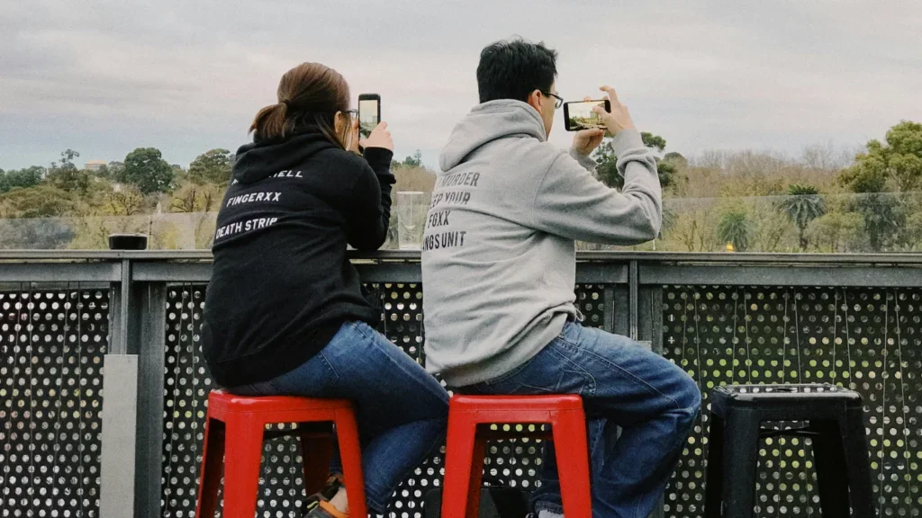 A couple wearing black and grey custom hoodies while filming something using their mobile phones.