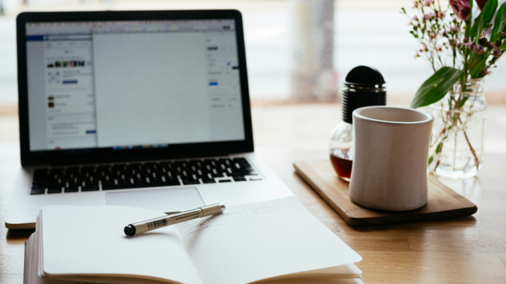A laptop and a notebook of a freelance copywriter on a desk in a cozy environment.