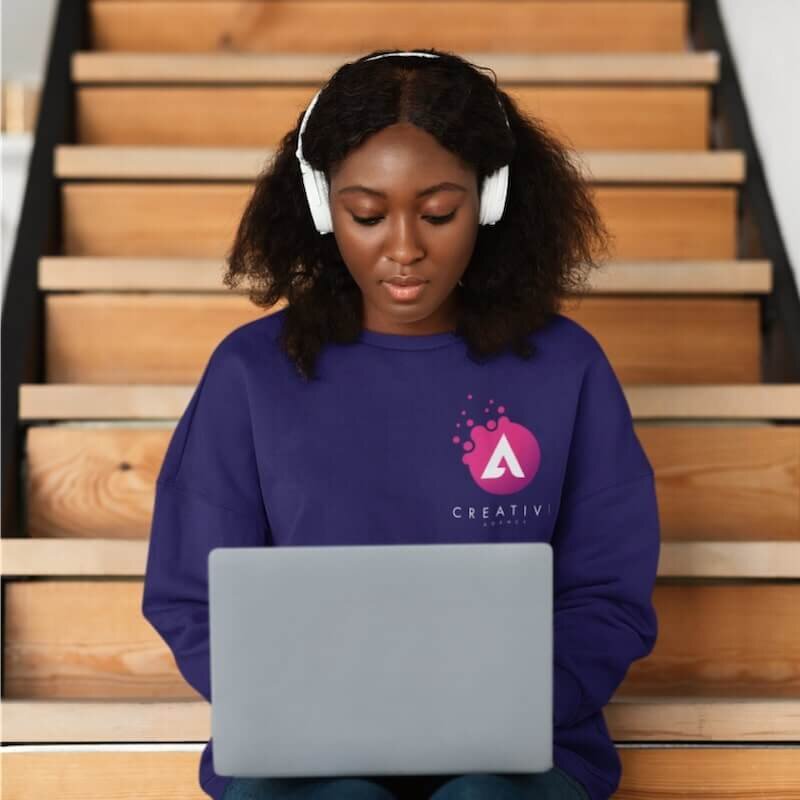 A woman working on a laptop wearing headphones and a purple custom hoodie with a company logo.