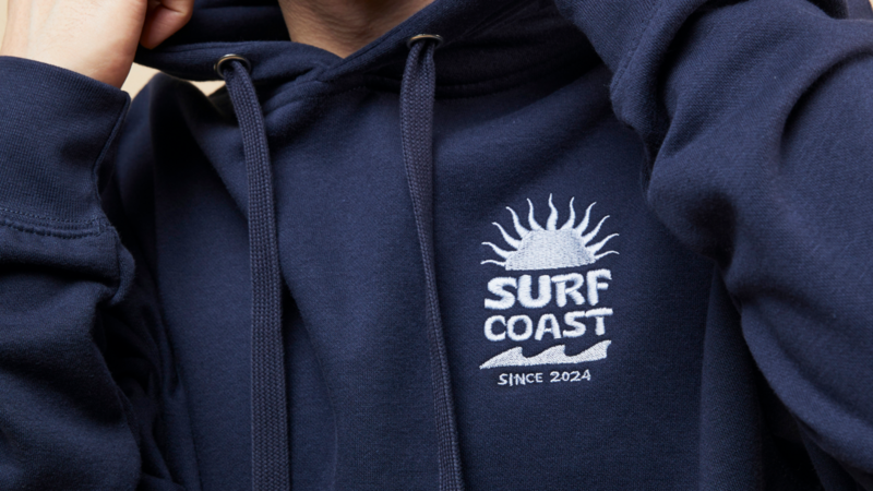 A person wearing a navy blue hoodie featuring a logo with the text "SURF COAST" and "SINCE 2024," along with a sun design and waves.