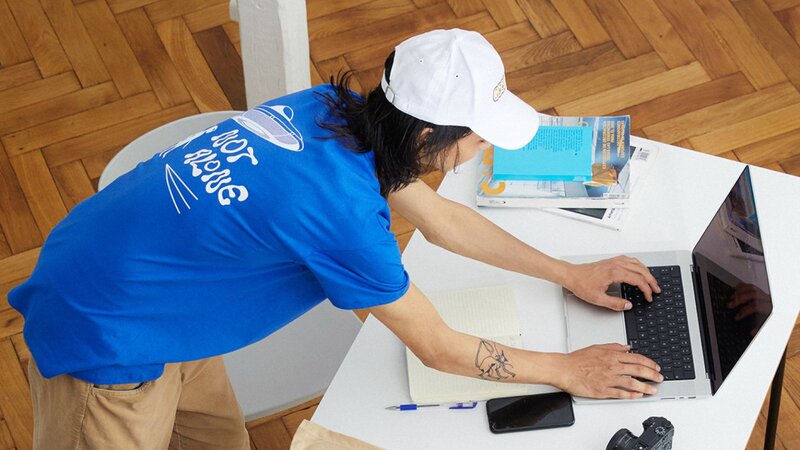 A man in a blue t-shirt and white cap is typing on a laptop. T-shirt, notebook and journals next to laptop (on top a table).