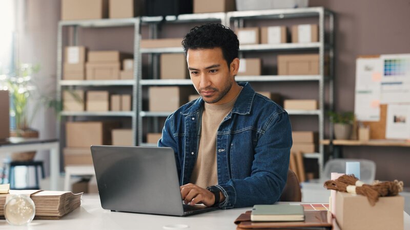 A man with gray hair sitting on a couch, looking thoughtfully at a laptop in a bright room.