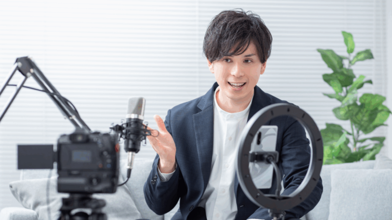 A man in a suit records a video with a camera, microphone, and ring light.
