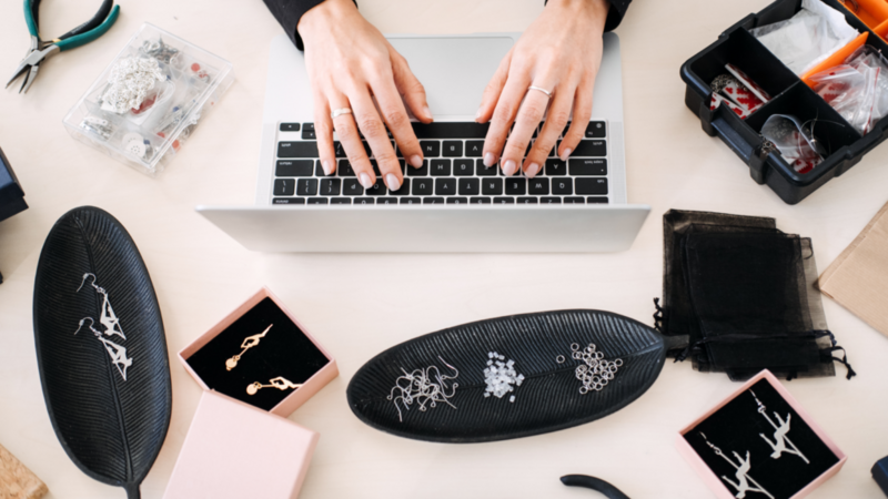 A woman sits at a table, using a laptop surrounded by an assortment of jewelry pieces.