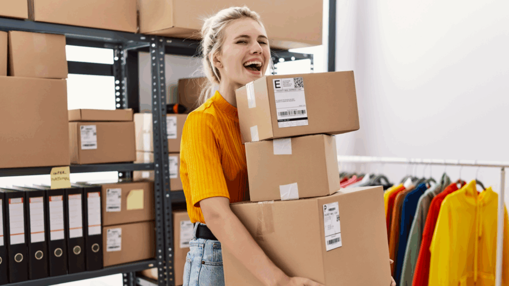 A woman holds several boxes while standing in a warehouse, surrounded by shelves and storage items.