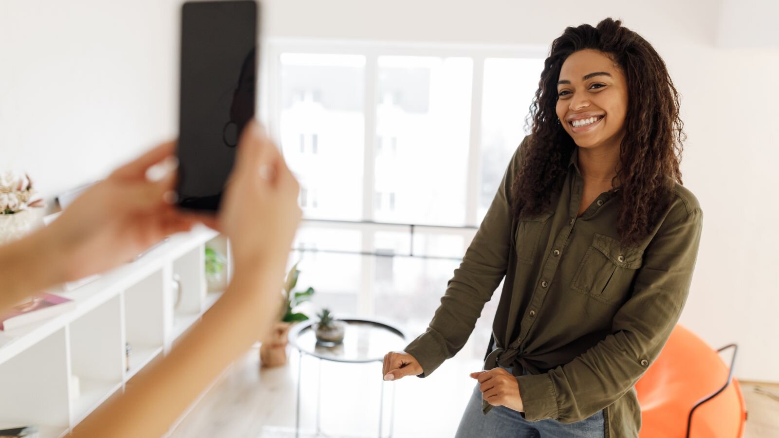 A woman stands indoors, smiling, as another person photographs her on a smartphone.