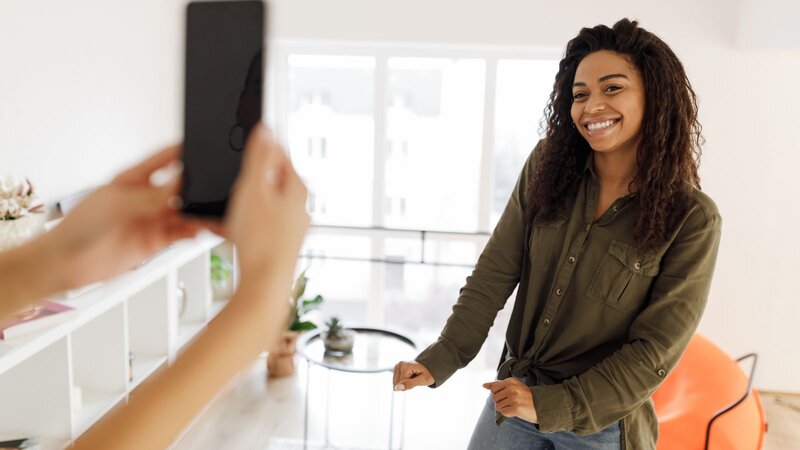 A woman stands indoors, smiling, as another person photographs her on a smartphone.