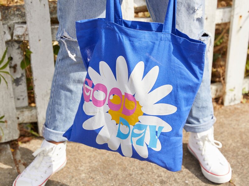 A person holding a blue tote bag with a floral design that includes the words "GOOD" and "DAY".