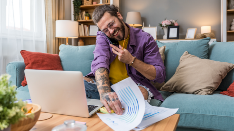 A cheerful man with tattoos talks on a phone, sitting on a cozy sofa. He's reviewing paperwork by a laptop.