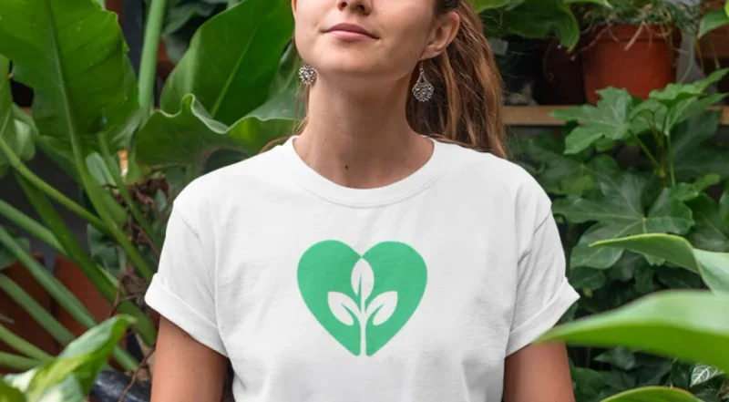 A woman in a green heart t-shirt stands in front of lush green plants, smiling and enjoying the natural surroundings.