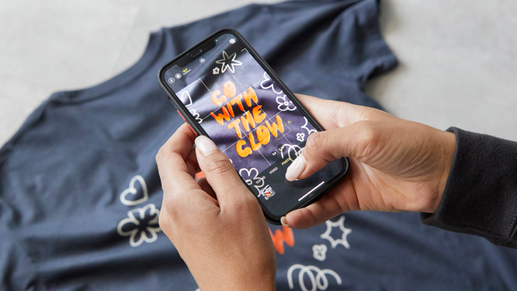A person is taking a picture of a blue “Go with the glow” t-shirt using a smartphone.