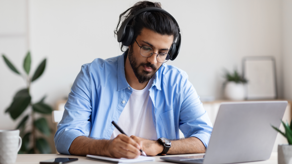 A man in a blue shirt wearing headphones, sitting at a desk with a laptop on it, writing something down.
