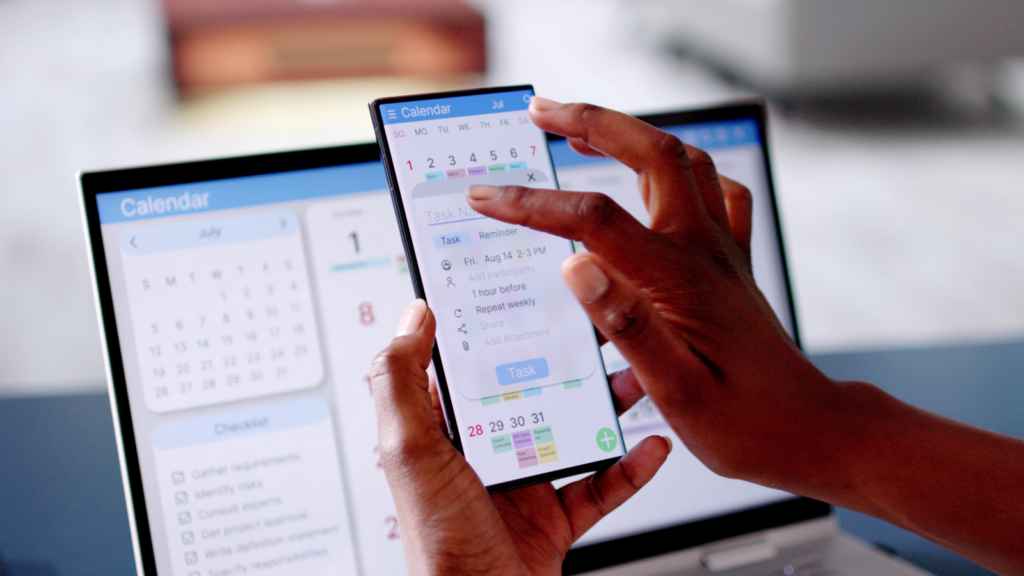 Hands holding a smartphone displaying a calendar app, inputting a task. A laptop in the background shows a calendar interface.